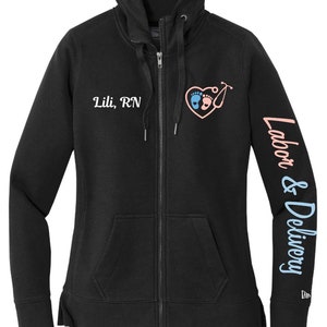 May include: Black zip-up hoodie with a white embroidered heart design featuring two footprints and a stethoscope. The text "Lili, RN" is embroidered on the left chest and "Labor & Delivery" is printed on the right sleeve.