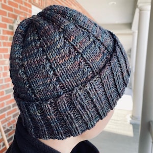 May include: A dark blue knitted beanie with a textured pattern. The beanie is made with a multi-colored yarn with shades of blue, purple, and brown.