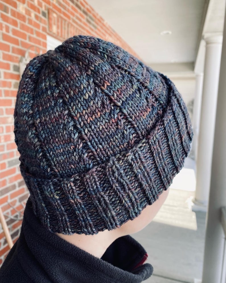 May include: A dark blue knitted beanie with a textured pattern. The beanie is made with a multi-colored yarn with shades of blue, purple, and brown.