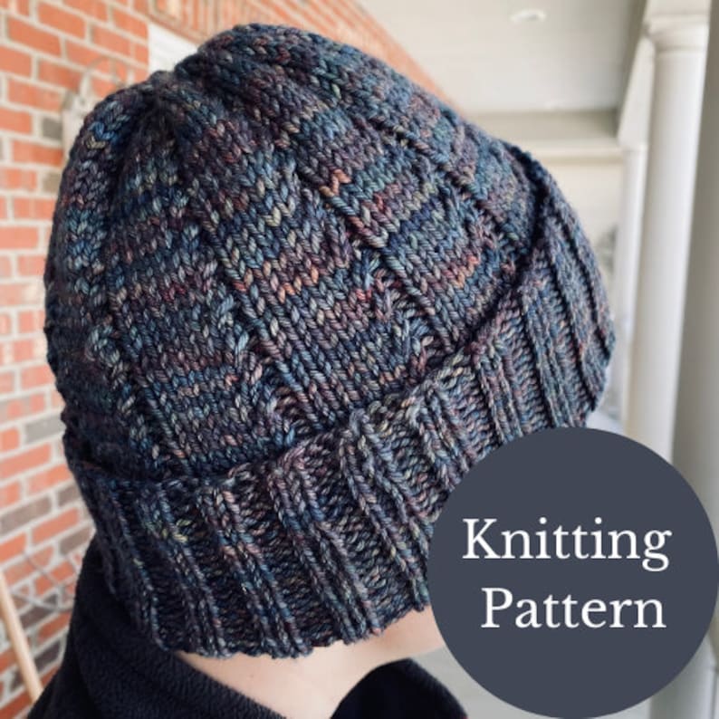 May include: A close-up of a dark blue and grey knitted beanie with a ribbed brim. The beanie is made with a variegated yarn and has a subtle texture. The text "Knitting Pattern" is visible in the lower right corner of the image.
