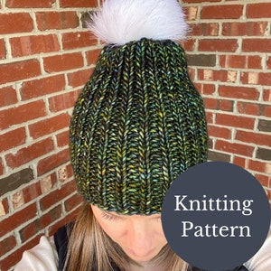 May include: A green and black knitted beanie with a white pom pom. The beanie has a ribbed texture and is made with a chunky yarn. The text "Knitting Pattern" is on a dark grey circle in the bottom right corner of the image.
