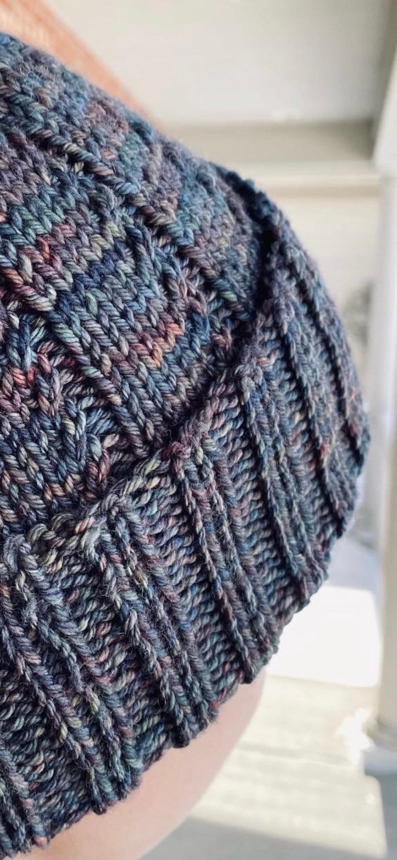 May include: A close-up of a knitted beanie hat in a dark blue, green, and purple color scheme. The hat has a cable knit pattern and a soft, fuzzy texture.
