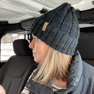 May include: A woman wearing a black and blue knit beanie with a brown leather tag that says "koal" on it. She is sitting in a car and wearing a black vest over a striped shirt.