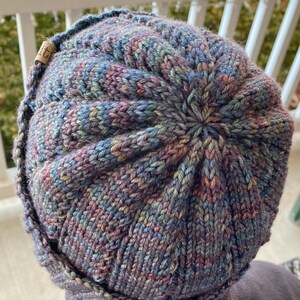 May include: A close-up of a knitted hat with a multi-colored yarn in shades of blue, purple, and green. The hat has a unique textured pattern and a small brown tag with the word "LIT" on it.