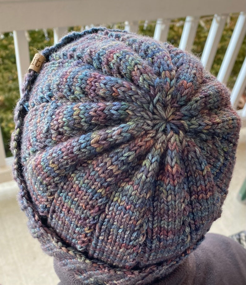 May include: A close-up of a knitted hat with a multi-colored yarn in shades of blue, purple, and green. The hat has a unique textured pattern and a small brown tag with the word "LIT" on it.