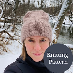 Op de afbeelding: Een close-up van een vrouw die een gebreide muts draagt in een lichtroze kleur. De muts is een eenvoudige beanie-stijl met een omslagrand. De vrouw staat in een besneeuwde buitenomgeving. De tekst "Knitting Pattern" is over de afbeelding gelegd.
