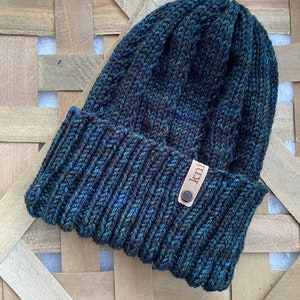 May include: A dark blue knitted beanie with a brown leather tag that says "knit". The beanie is sitting on a woven basket.