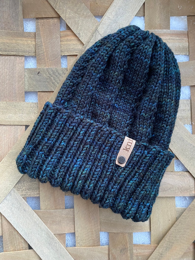 May include: A dark blue knitted beanie with a brown leather tag that says "knit". The beanie is sitting on a woven basket.