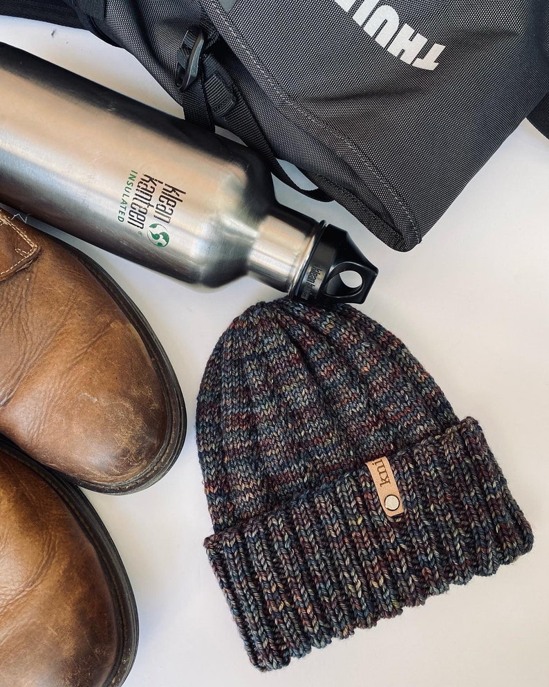 May include: A brown leather boot, a silver insulated water bottle with the text "Klean Kanteen Insulated", and a dark blue and brown knit beanie with a brown leather tag that says "KULL" on a white surface.