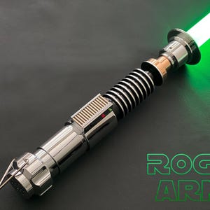 May include: A metallic lightsaber prop with a glowing green blade, featuring a silver handle with black ribbed grip sections, a copper-colored neck, and a rectangular control box with small red and green lights. The text ROGUE ARMS is visible.