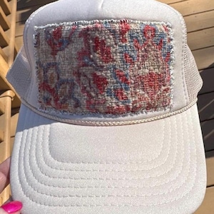 Puede incluir: Gorra de camionero blanca con parte trasera de malla beige y un parche decorativo. El parche presenta un diseño floral colorido en rojo, azul y beige. La gorra tiene una visera curva y un botón blanco en la parte superior.