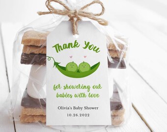 Op de afbeelding: Bedankt voor het vieren van onze baby's met liefde. Olivia's babyshower 10.26.2022. Een wit label met groene erwten in een peul en roze hartjes. Het label is vastgemaakt aan een doorzichtige plastic zak met een stapel s'mores erin.