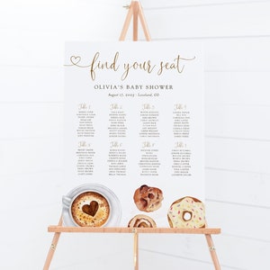 May include: A white seating chart with gold text for a baby shower. The chart is titled "Find Your Seat" and lists guests by table number. The chart is decorated with illustrations of coffee, donuts, and pastries.