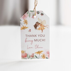 Pode incluir: Uma etiqueta de agradecimento com uma ilustra&ccedil;&atilde;o em aquarela de uma fada com cabelo castanho e um vestido amarelo voando sobre um campo de flores. O texto na etiqueta diz "Obrigado fada! Com amor, Olivia."
