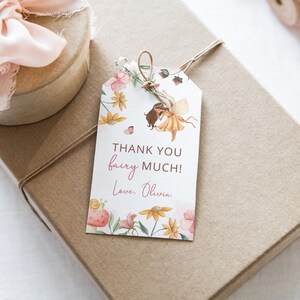 Pode incluir: Uma etiqueta de presente branca com uma ilustra&ccedil;&atilde;o em aquarela de uma fada com flores e borboletas. O texto na etiqueta diz "Obrigado fada muito! Com amor, Olivia."