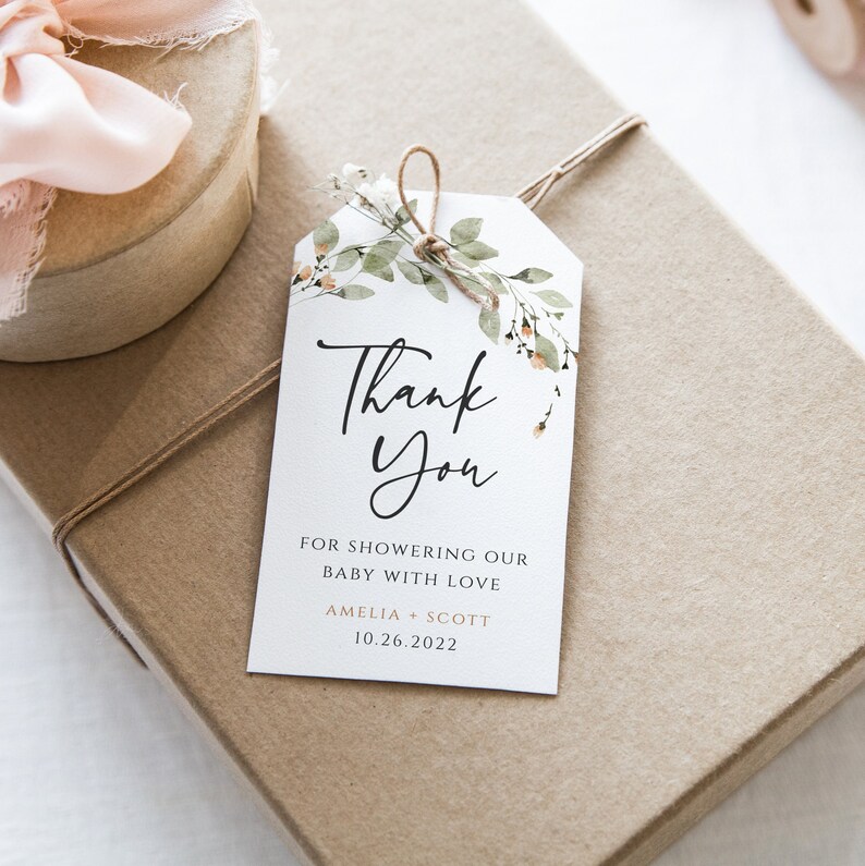May include: A white thank you tag with a floral design in shades of green and peach. The tag says "Thank You" in black script font and "For showering our baby with love Amelia + Scott 10.26.2022" in black font.