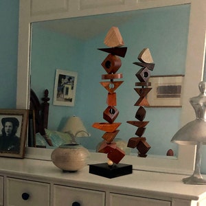 May include: Two wooden sculptures made of stacked geometric shapes in various shades of brown and red. The sculptures are displayed on a black base and are reflected in a mirror behind them. The sculptures are on a white dresser with drawers.