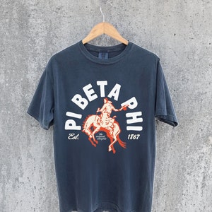 Könnte beinhalten: Ein dunkelblaues T-Shirt mit einer weißen Grafik einer Person, die ein Pferd reitet. Die Grafik hat die Form eines Kreises mit dem Text "Pi Beta Phi" um den Außenrand. Der Text "Est. 1867" befindet sich unterhalb der Grafik.