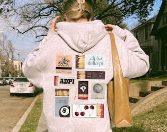 May include: A gray hooded sweatshirt with a variety of colorful patches on the back. The patches include a cowboy riding a horse, a matchbook with the text "ADPI", a matchbook with the text "alpha delta pi", a matchbook with the text "lucky me to be a girl", a matchbook with the text "18", a matchbook with the text "51", a moon, a 8-ball, and cherries.