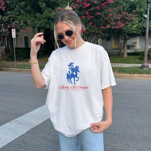 Può includere: Una maglietta bianca con una grafica blu e rossa di una persona a cavallo. La grafica include il testo "Alpha Chi Omega".