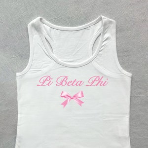 May include: White tank top with a pink bow and the text "Pi Beta Phi" in pink cursive lettering.