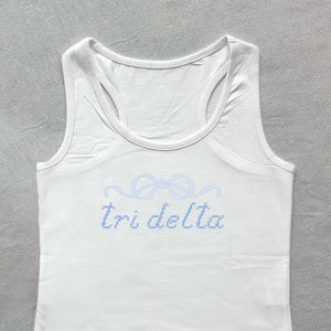 Puede incluir: Camiseta blanca de tirantes con un lazo bordado en azul y el texto "Tri Delta" en la parte delantera.