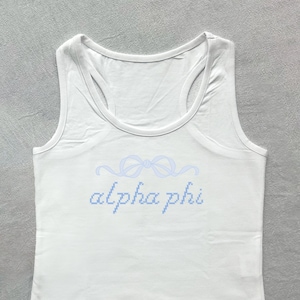 Puede incluir: Camiseta blanca de tirantes con un diseño de cinta azul claro y el texto "alpha phi" en azul claro.