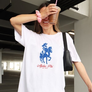 Op de afbeelding: Wit T-shirt met een blauwe en rode afbeelding van een vrouw die paardrijdt. De afbeelding bevat de tekst "Alpha Phi est. 1872".