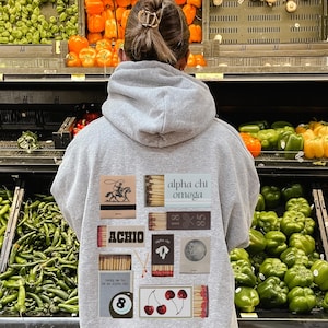 Può includere: Una felpa con cappuccio grigia con una varietà di toppe colorate che raffigurano fiammiferi, una palla da biliardo, ciliegie e testo che dice "alpha chi omega" e "ACHIO".