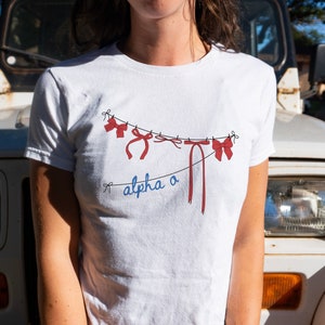 Könnte beinhalten: Weißes T-Shirt mit einer roten und blauen Grafik von Schleifen, die an einer Wäscheleine hängen, mit dem Text "alpha o" darunter.