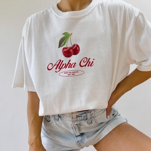 Può includere: Maglietta bianca con una grafica di ciliegie rosse e il testo "Alpha Chi" in corsivo rosso. Sotto il testo c'è un ovale rosso con il testo "Seek the Heights Est. 1988".