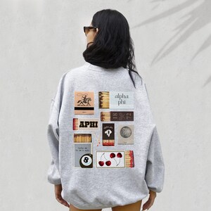 May include: Grey sweatshirt with a graphic design of matchboxes featuring the text "APHI" and "alpha phi". The design also includes a cowboy riding a horse, cherries, and a pool ball.