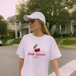 May include: A white t-shirt with a red cherry graphic and the text "Delta Gamma" in red. The shirt is worn by a person with blonde hair and sunglasses.