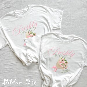 May include: Two white t-shirts with the text "Freshly Picked" in pink and gray. One shirt has the text "Little" below the main text. Both shirts feature a bouquet of pink flowers.
