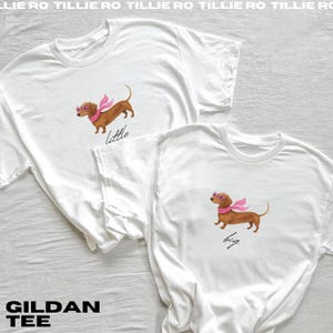 May include: Two white Gildan tee shirts. One shirt has a watercolour illustration of a brown dachshund wearing pink sunglasses and a pink scarf with the word "little" below. The other shirt has the same illustration with the word "big".