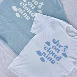 May include: Two t-shirts with the text "she's on cloud nine" printed in blue on a white background. The shirts are laid out on a white surface.