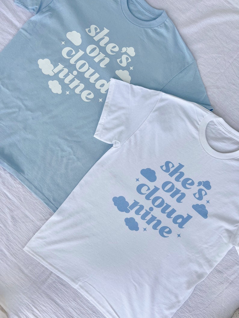 May include: Two t-shirts with the text "she's on cloud nine" printed in blue on a white background. The shirts are laid out on a white surface.