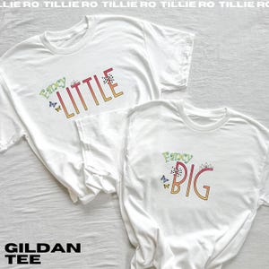 Pode incluir: Duas camisetas Gildan brancas com as palavras "Fancy Little" e "Fancy Big" em letras coloridas e estilizadas. Borboletas e detalhes florais decoram o texto. As camisetas s&atilde;o roupas casuais.