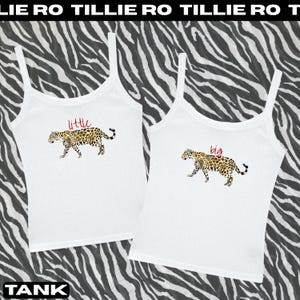 May include: Two white tank tops with spaghetti straps. One top has the word "little" in red above a leopard illustration. The other top has the word "big" in red above a leopard illustration. The background is a black and white zebra print.