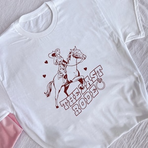 May include: White t-shirt with a brown illustration of a woman riding a horse. The text "THE LAST RODEO" is written in brown with a diamond ring above the word "RODEO".