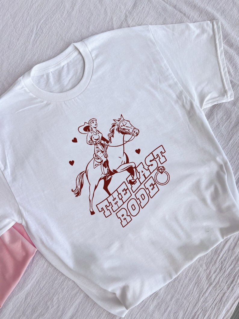 May include: White t-shirt with a brown illustration of a woman riding a horse. The text "THE LAST RODEO" is written in brown with a diamond ring above the word "RODEO".