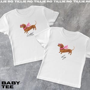 May include: Two white baby tees featuring a watercolor dachshund wearing pink sunglasses and a scarf. One shirt says "little" and the other says "big". The text "BABY TEE" is at the bottom left.
