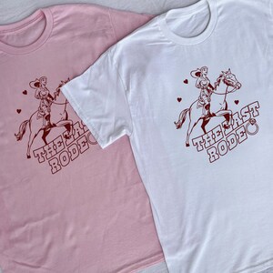May include: Two t-shirts with a graphic of a woman riding a horse. The text on the shirts reads "The Last Rodeo". One shirt is pink and the other is white.