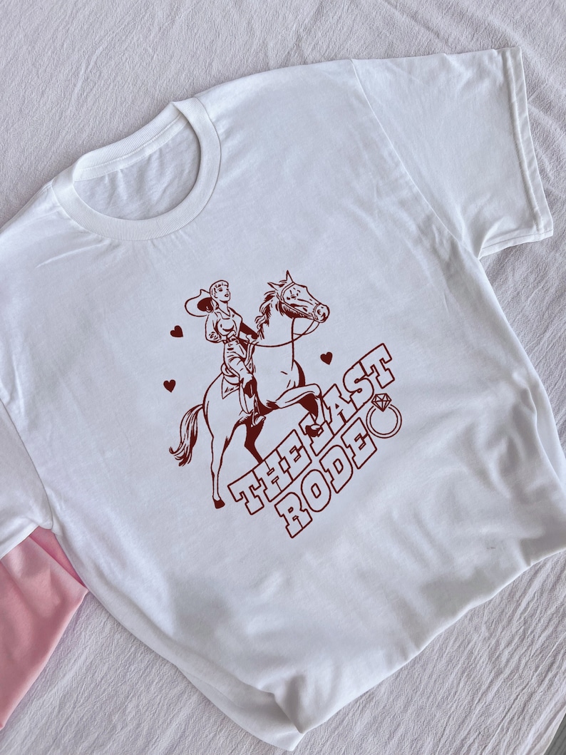 May include: White t-shirt with a brown illustration of a woman riding a horse. The text "THE LAST RODEO" is printed on the shirt with a diamond ring symbol.