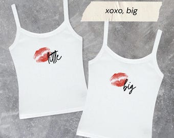 Könnte beinhalten: Zwei weiße Tanktops mit Spaghettiträgern. Eines hat einen roten Lippenabdruck und das Wort "little" in schwarzer Schrift. Das andere hat einen roten Lippenabdruck und das Wort "big" in schwarzer Schrift. Der Text "xoxo, big" steht über dem zweiten Top.