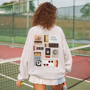 Może przedstawiać: Szara bluza z różnymi kolorowymi naszywkami, w tym pudełkiem zapałek z napisem "Axid", pudełkiem zapałek z napisem "alpha xi delta", pudełkiem zapałek z wiśniami i pudełkiem zapałek z bilą.