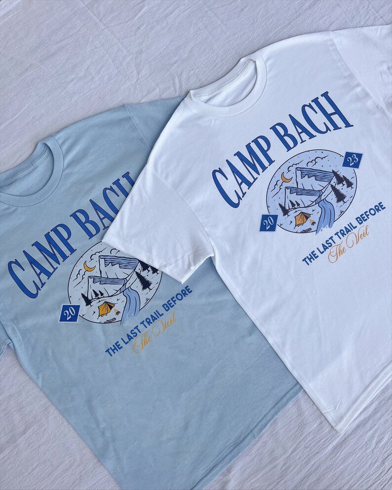 Op de afbeelding: Twee lichtblauwe en witte T-shirts met een grafisch ontwerp van een berglandschap en de tekst "CAMP BACH" en "THE LAST TRAIL BEFORE The Veil".