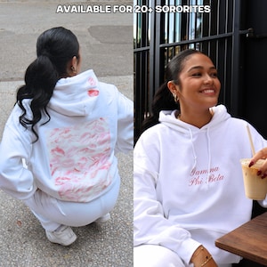 Op de afbeelding: Twee vrouwen dragen witte hoodies. De ene hoodie heeft een roze en witte print van lippen op de rug. De andere hoodie heeft de tekst "Gamma Phi Beta" in roze op de voorkant.