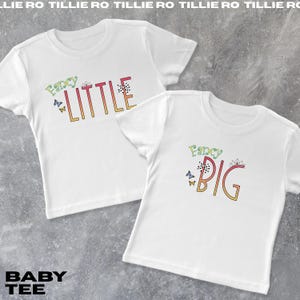 Pode incluir: Duas camisetas brancas para beb&ecirc;s com as palavras "Fancy Little" e "Fancy Big" em letras coloridas. As camisas t&ecirc;m gr&aacute;ficos de borboletas e coroas. O texto "BABY TEE" est&aacute; na parte inferior.