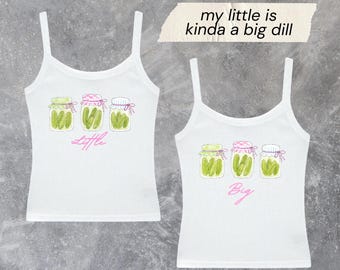 May include: Two white tank tops featuring a graphic of three glass jars filled with green pickles. The left tank top has the word "Little" in pink script, and the right tank top has the word "Big" in pink script. A piece of tape displays "my little is kinda a big dill".